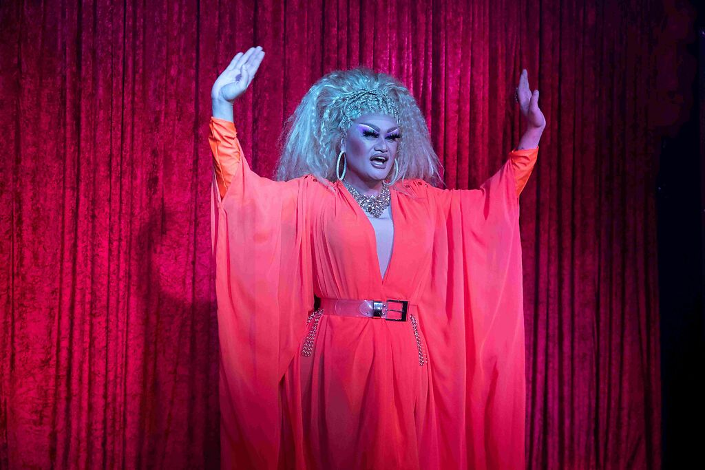 Drag queen on stage in front of a red curtain, dressed in a flowing red gown, holding their hands in the air
