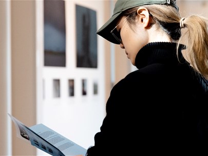 A person with a ponytail dressed in a black jumper and a cap reading a booklet with artwork in the background
