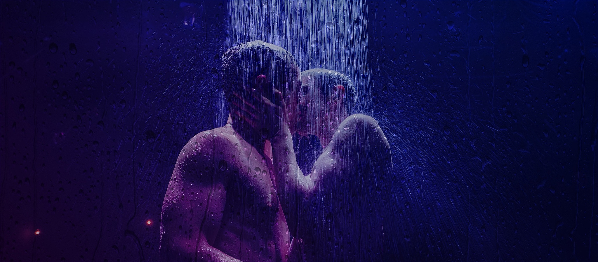 Two shirtless men embrace each other under stage lights and a shower of water.
