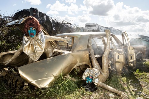 Two people with green faces and gold costumes in front of a gold-coloured car wreck