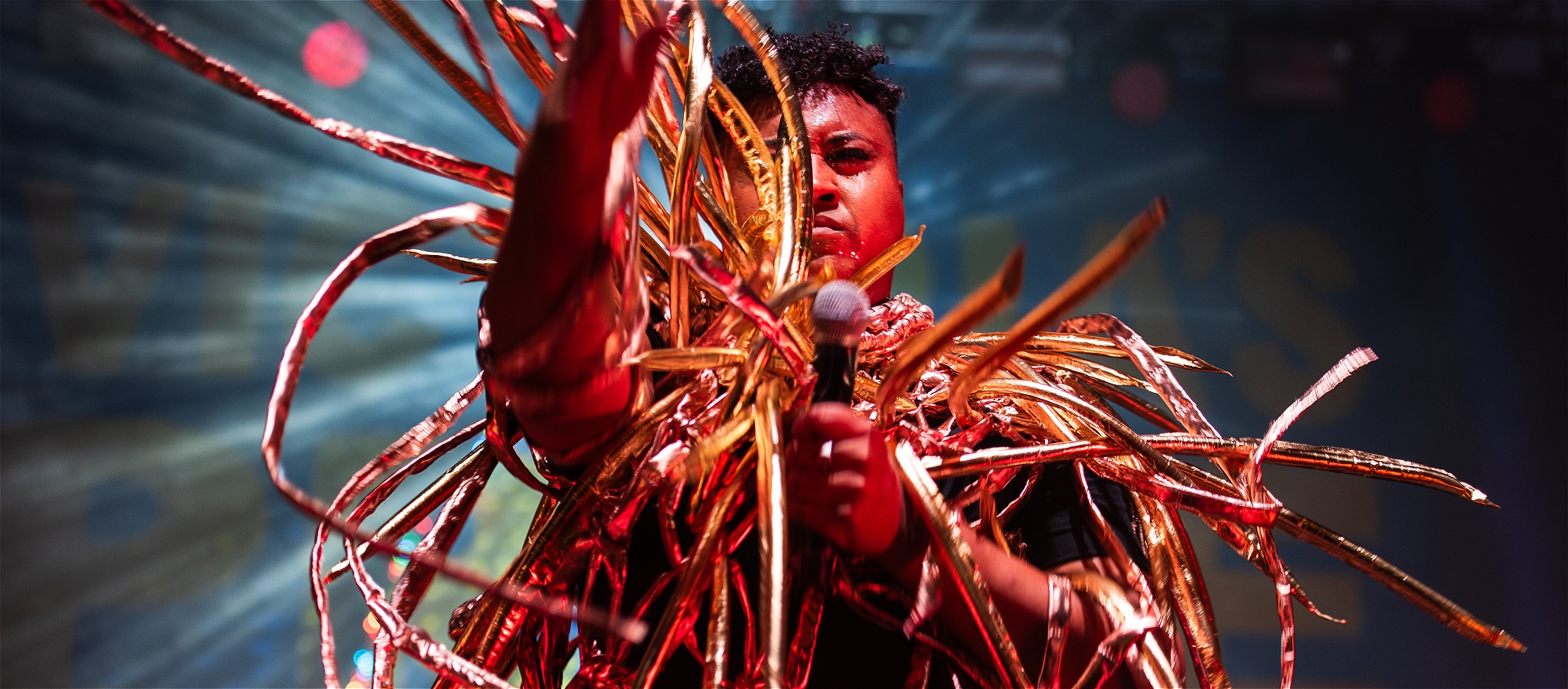 A BIPOC artist dancing on stage holding a microphone. They are wearing a gold metallic costume that has tentacles joyfully flying in every direction.