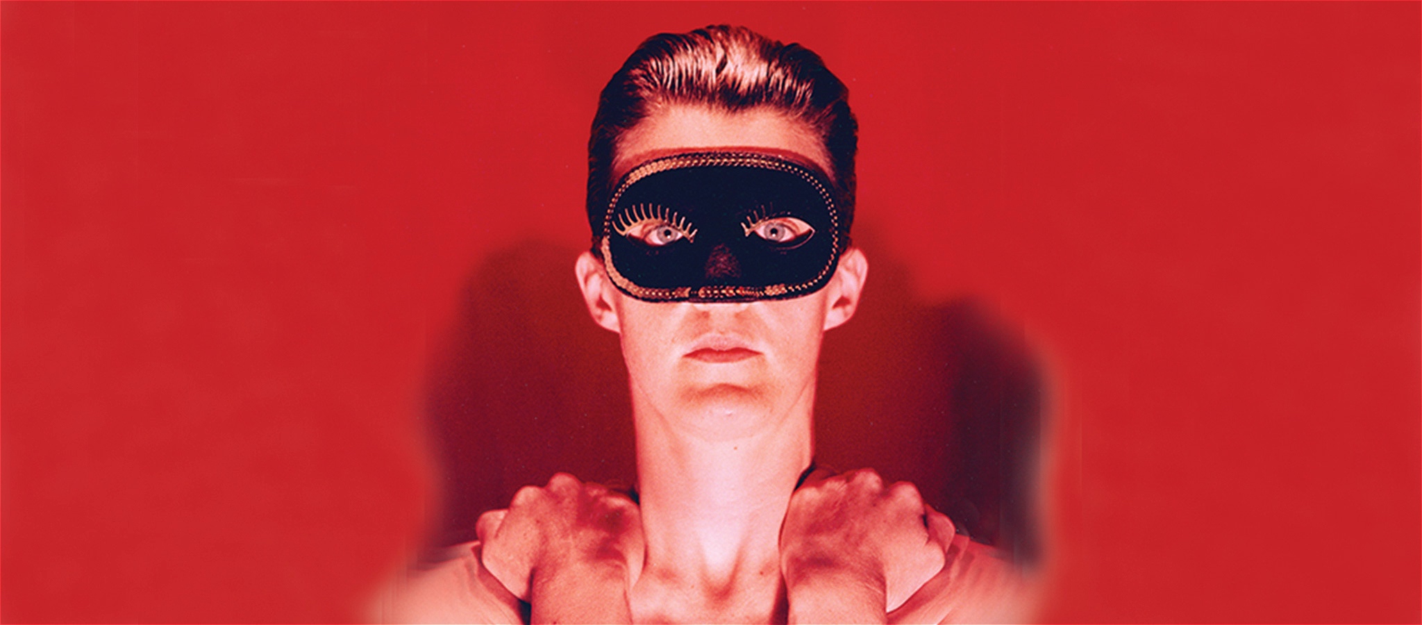A non gender specific person wearing a black mask and with short slick back hair stares back at the viewer in a red background.