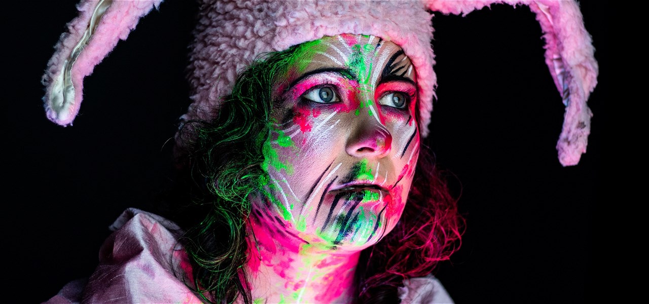 A woman is staring pensively in the distance. She is covered in florescent face paint and wearing pink bunny ears.