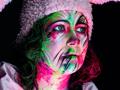A woman is staring pensively in the distance. She is covered in florescent face paint and wearing pink bunny ears.