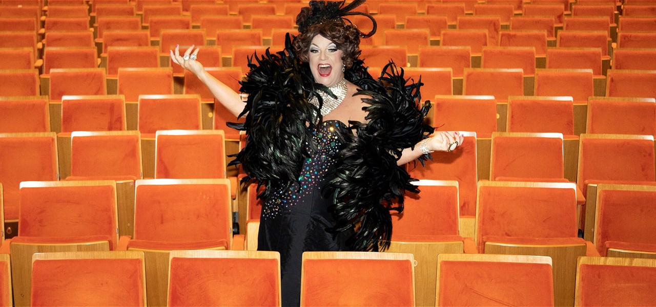 Dolly stands in the seats of a large theatre. She is smiling, mouth open, hands in the air, and wearing a black gown.
