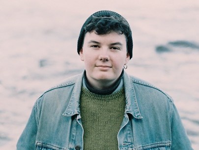 Sar is gazing into the camera while wearing a knitted beanie, a denim jacket and multiple layers of jumpers. They have short dark hair and one earring.