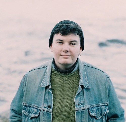 Sar is gazing into the camera while wearing a knitted beanie, a denim jacket and multiple layers of jumpers. They have short dark hair and one earring.