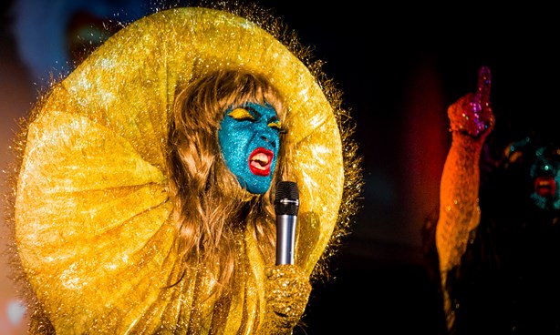 Person in gold wig with a massive gold headdress, face painted glittery blue, singing with one arm raised in defiance
