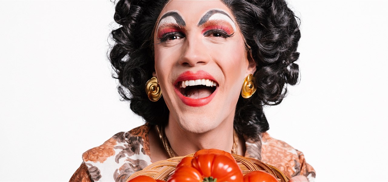 A heavily made-up drag queen, dressed as a Nonna, holding a bowl of large, red tomatoes.