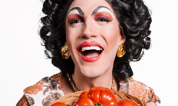 A heavily made-up drag queen, dressed as a Nonna, holding a bowl of large, red tomatoes.