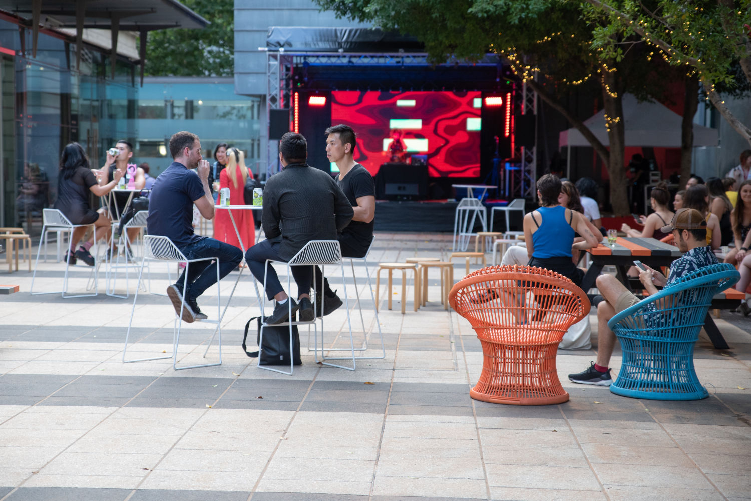 People sitting at socially-distanced tables in a courtyard, with an empty stage in the background