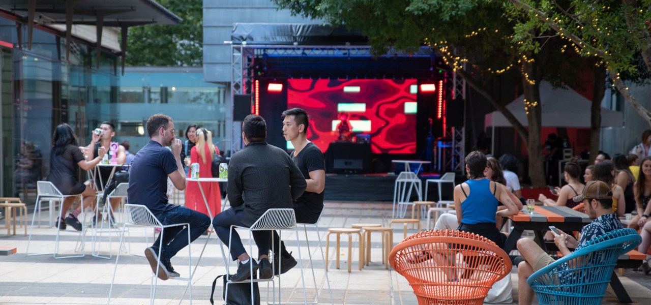People sitting at socially-distanced tables in a courtyard, with an empty stage in the background