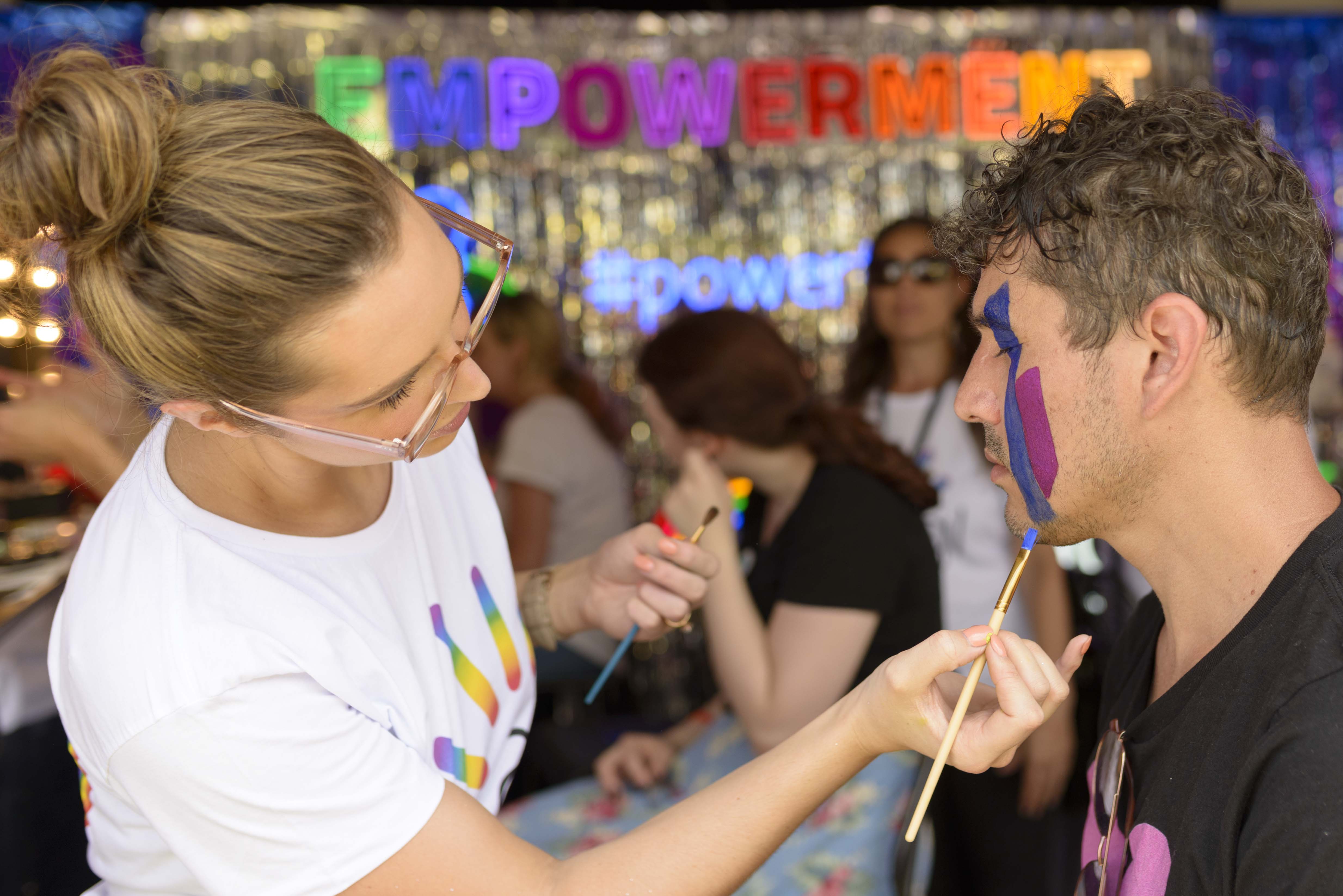 Young woman painting the face of a young man. Others and a banner reading "EMPOWERMENT"  in background