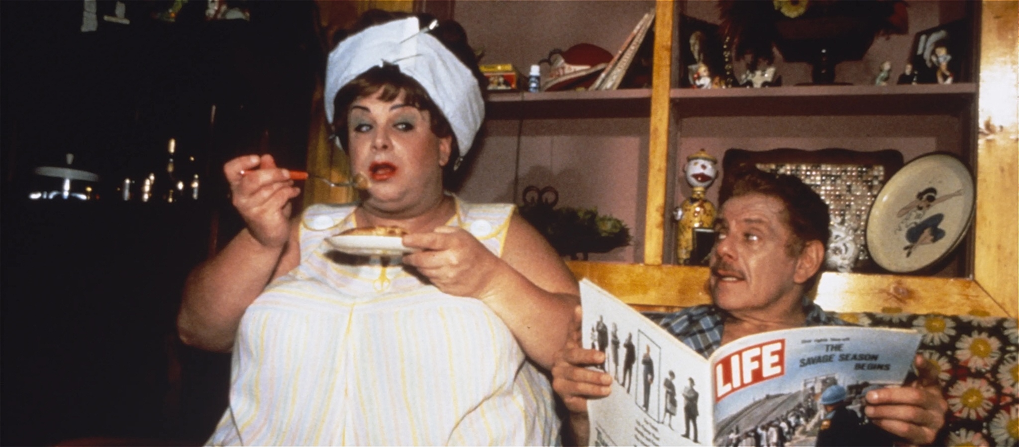 Still from Hairspray with Edna Turnblad eating from a plate and Wilbar Turnblad staring at her from behind a Life magazine