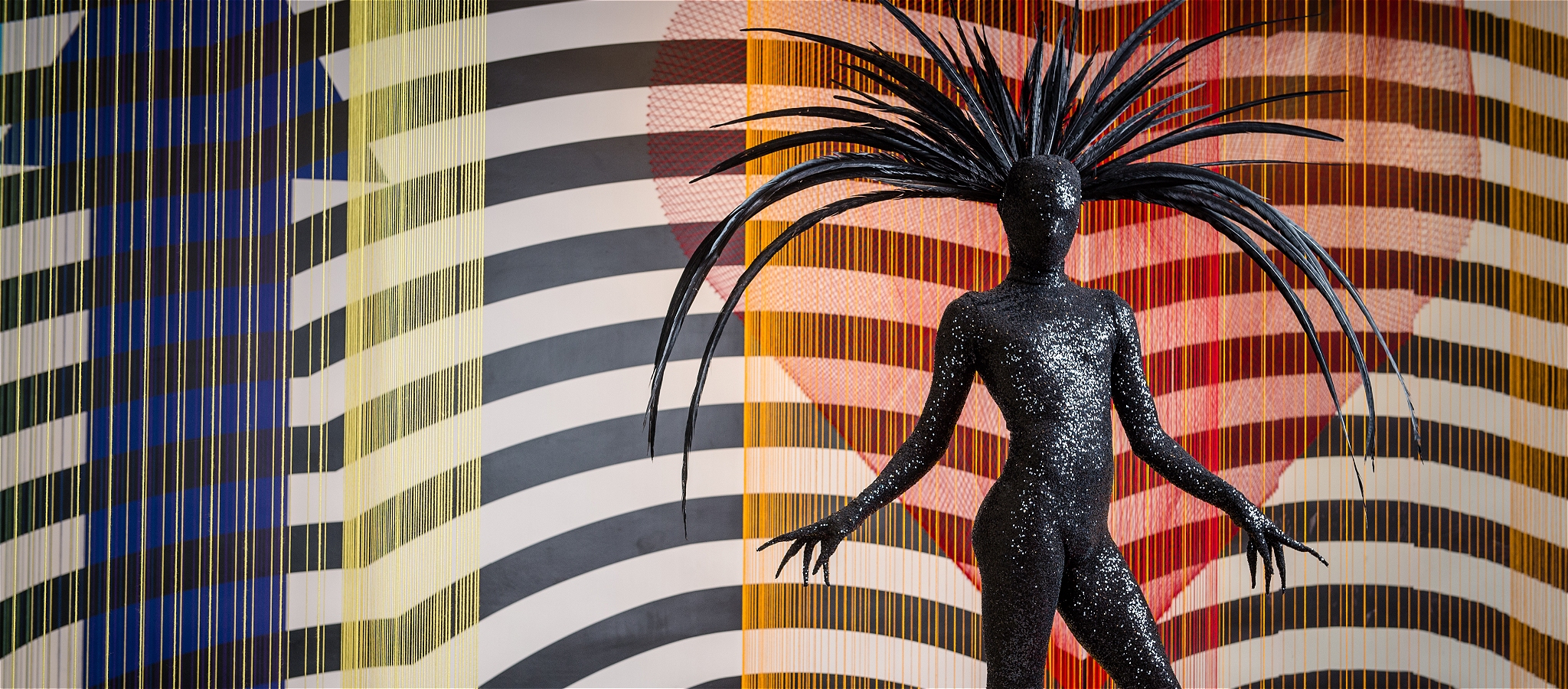 Person in a close-fitting black costume with extremely long fingernails and long spiky headress against a striped background