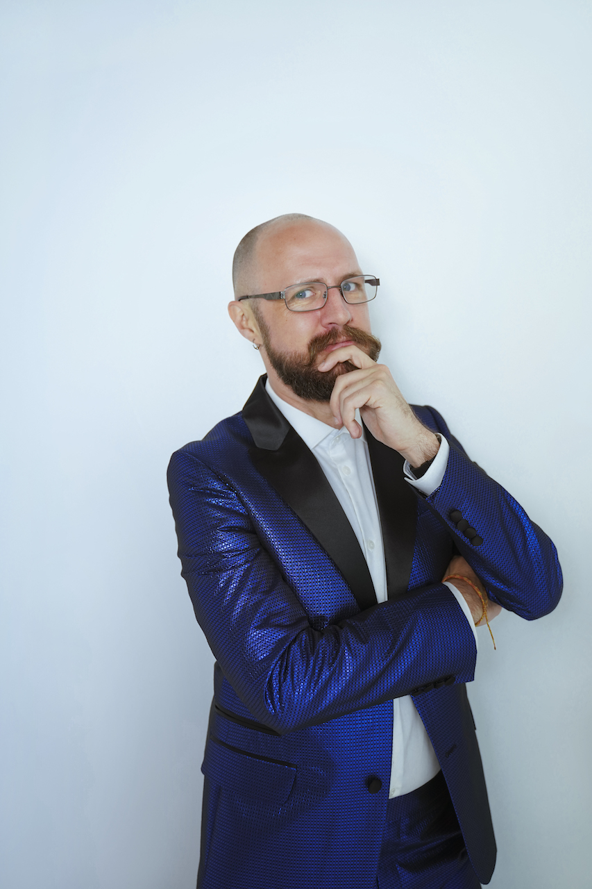 A person with a beard and glasses, wearing a blue suit with their hand on their chin.