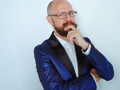 A person with a beard and glasses, wearing a blue suit with their hand on their chin.