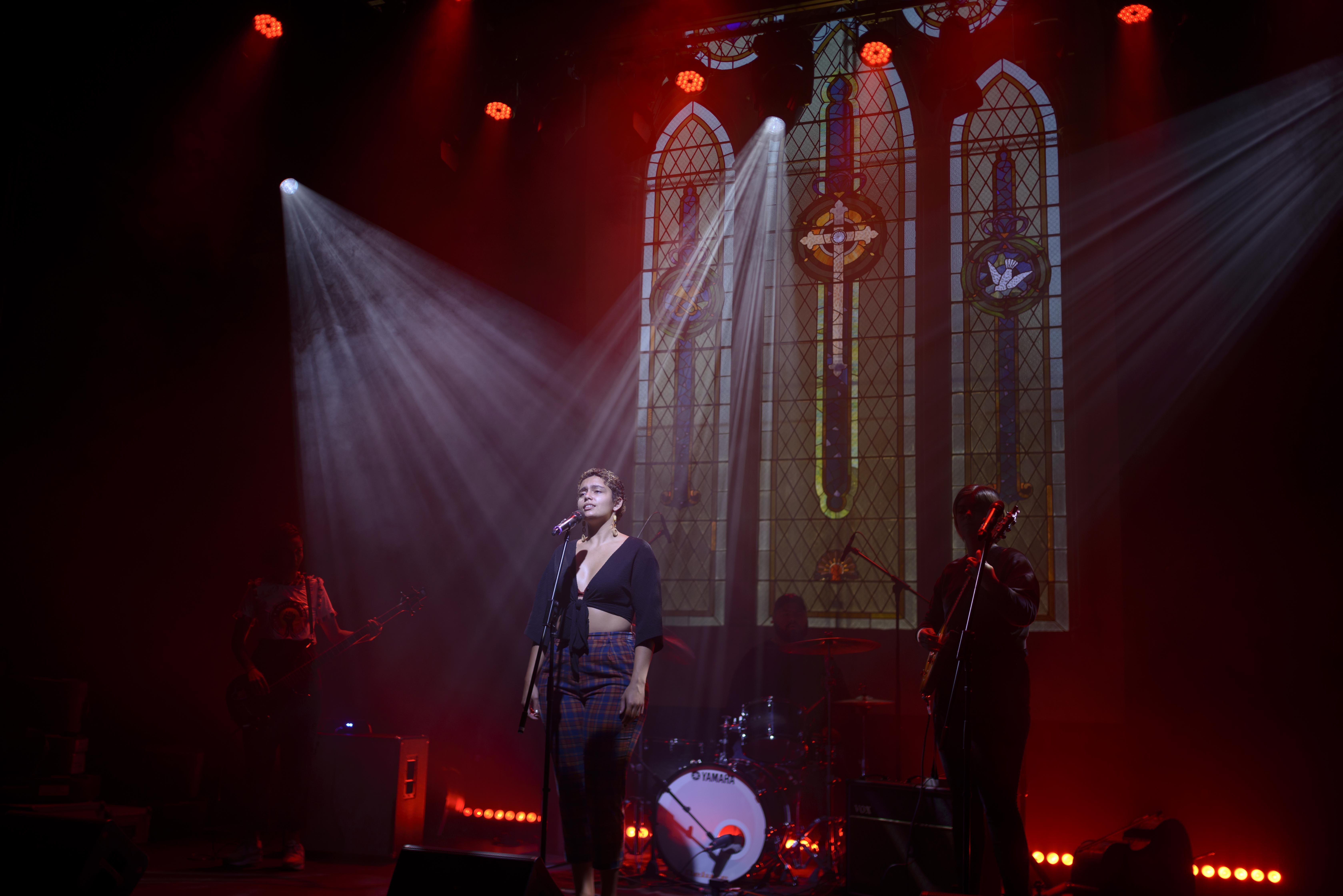 Singer on stage singing into a mic, stained glass windows behind, lights streaming down 'in a holy manner'