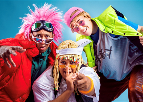 Group of three drag king performers gather together behind a blue background. The performer to the left wears a pink spiked wig and sunglasses, the performer next to them in the middle wears a blonde beaded wig and orange circle glasses and the performer on the furthest to the right wears a backwards pink baseball cap and neon green vest jacket