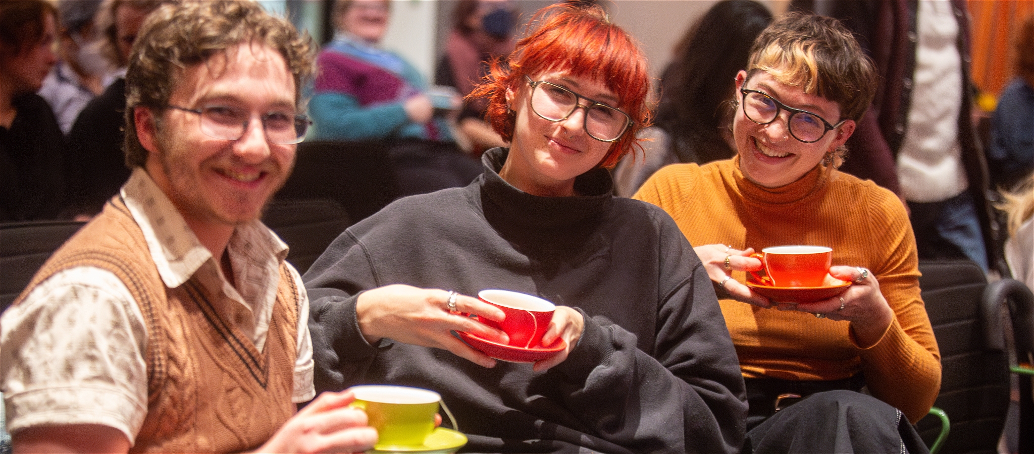 A photo showing three smiling people all wearing glasses and holding a cup of tea