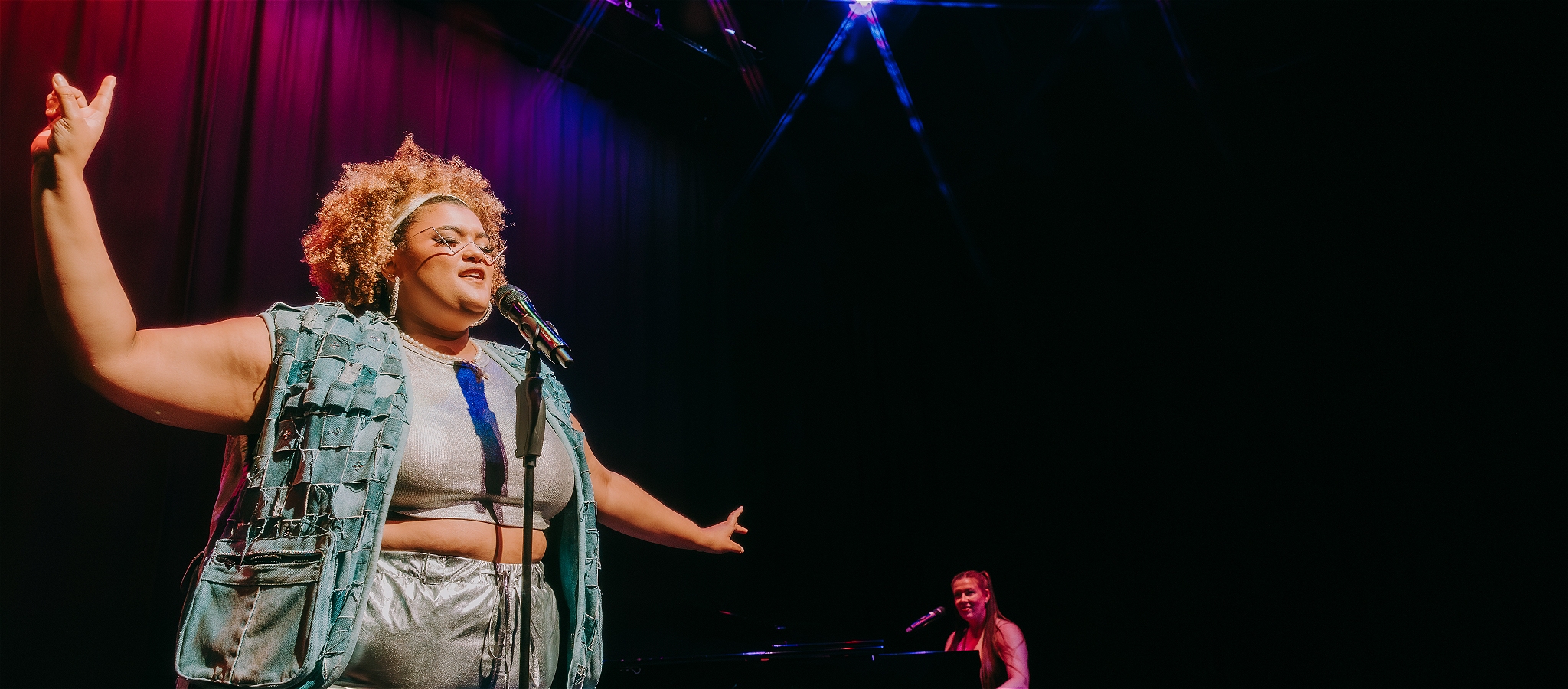  Milo singing live with arms outstretched at a microphone with accompanist on piano in the background
