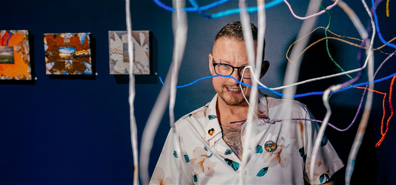 A white bespectacled trans man smiles at the camera wearing a white shirt. He is peering through a hanging sculpture. There are paintings behind him.