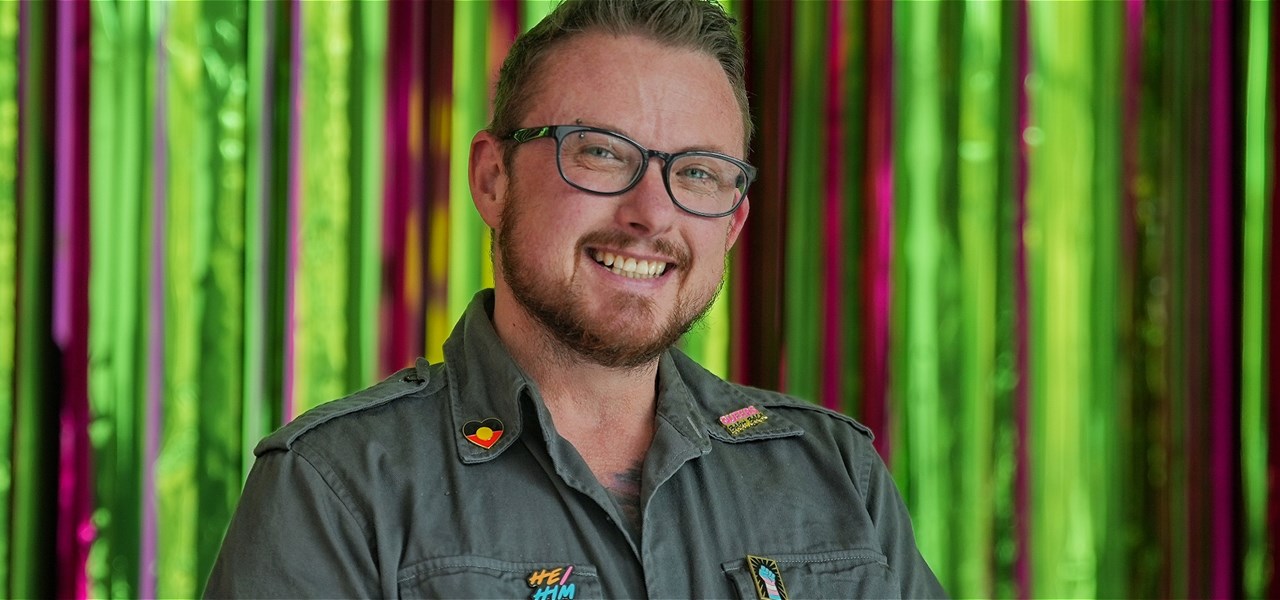 A white bespectacled trans man smiles at the camera wearing a khaki shirt with an assortment of badges pinned to it.