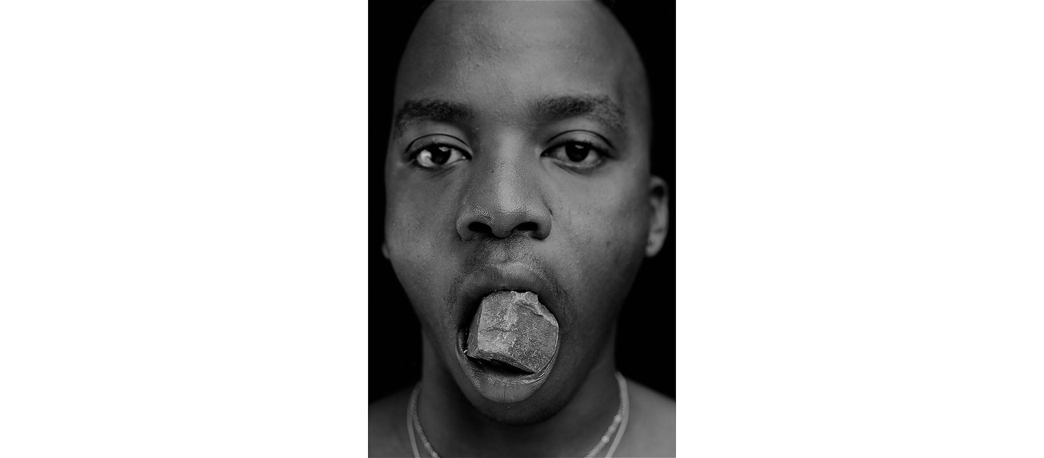 A black and white photograph of a person with a rock in their mouth, looking at the camera.