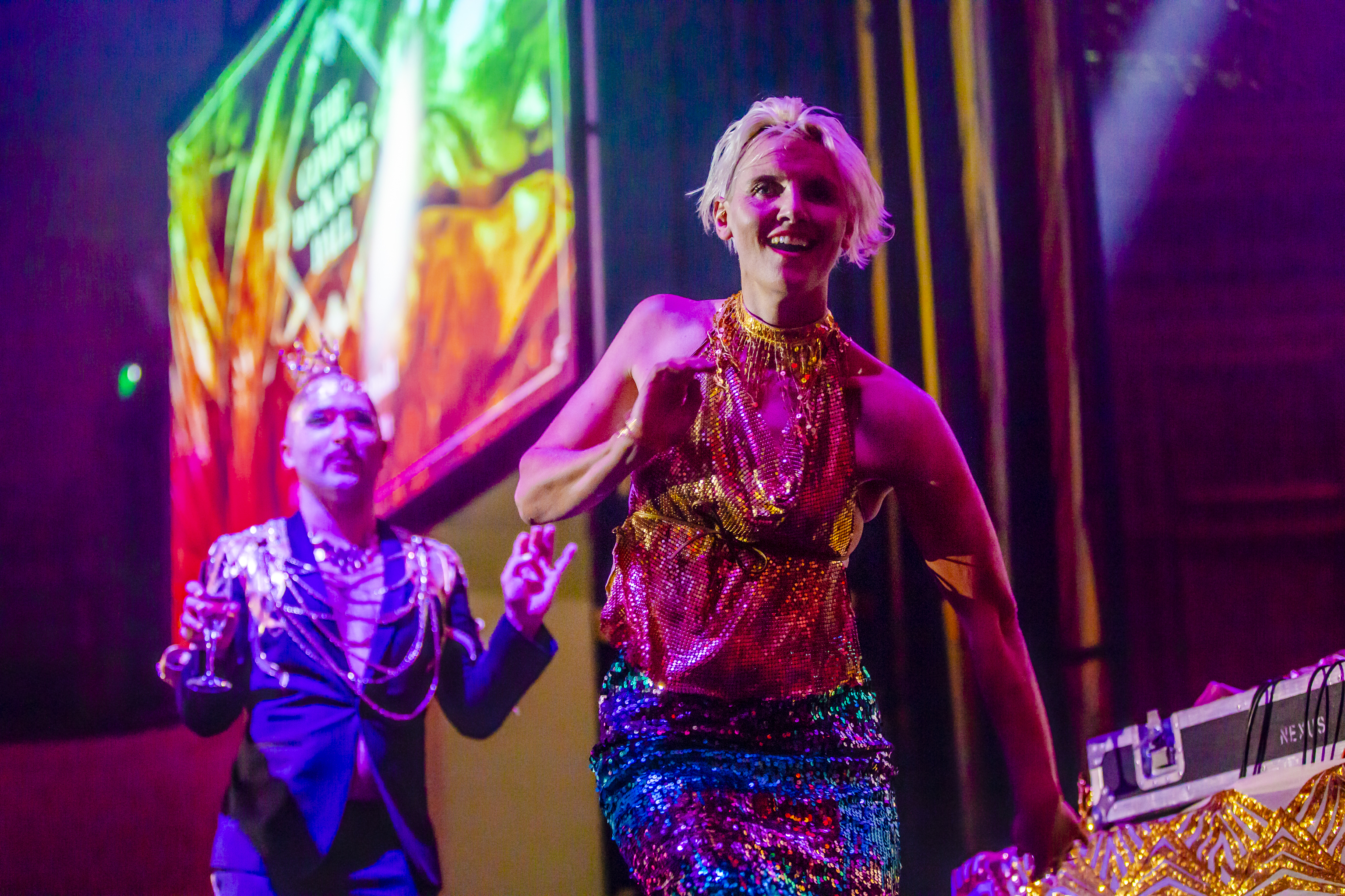 A woman wearing a bright sequin outfit and man dressed in a suit with a lot of jewellery are dancing toward camera.