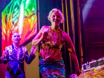 A woman wearing a bright sequin outfit and man dressed in a suit with a lot of jewellery are dancing toward camera.