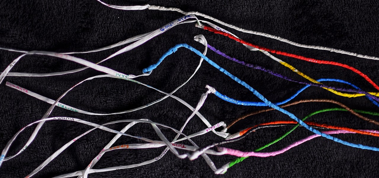 11 painted strands of raffia wrapped wire rest on a black background. On one end, white wavy strands of raffia have colourful words written on them.