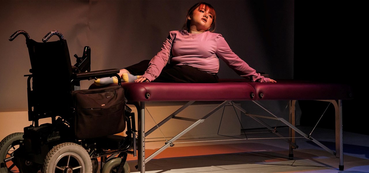 A girl in a pink jumper sits on a massage table looking out into the audience. Her wheelchair is next to the table.