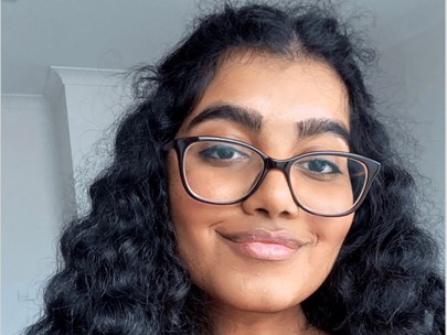 Purnima is wearing square framed glasses that have slipped down her nose. She smiles warmly and has thick, black, curly hair and defined eyebrows. They have brown skin and wear a red top.