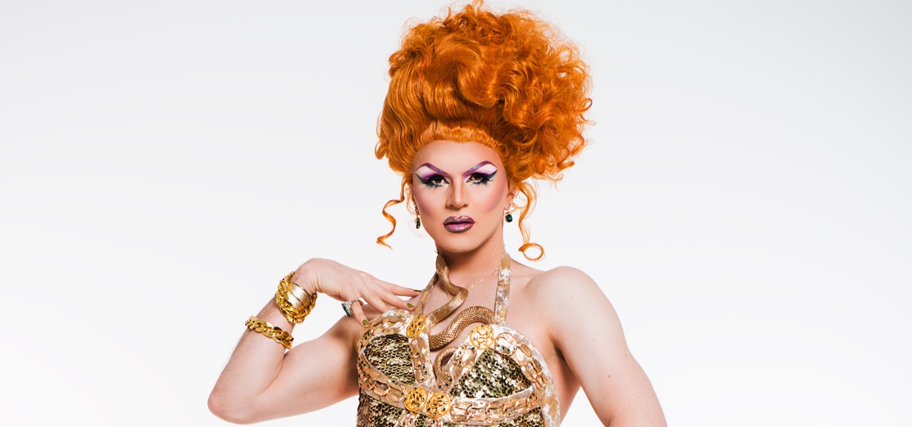 Valerie Hex in an orange wig and glamorous "metallic-like" dress; light grey background