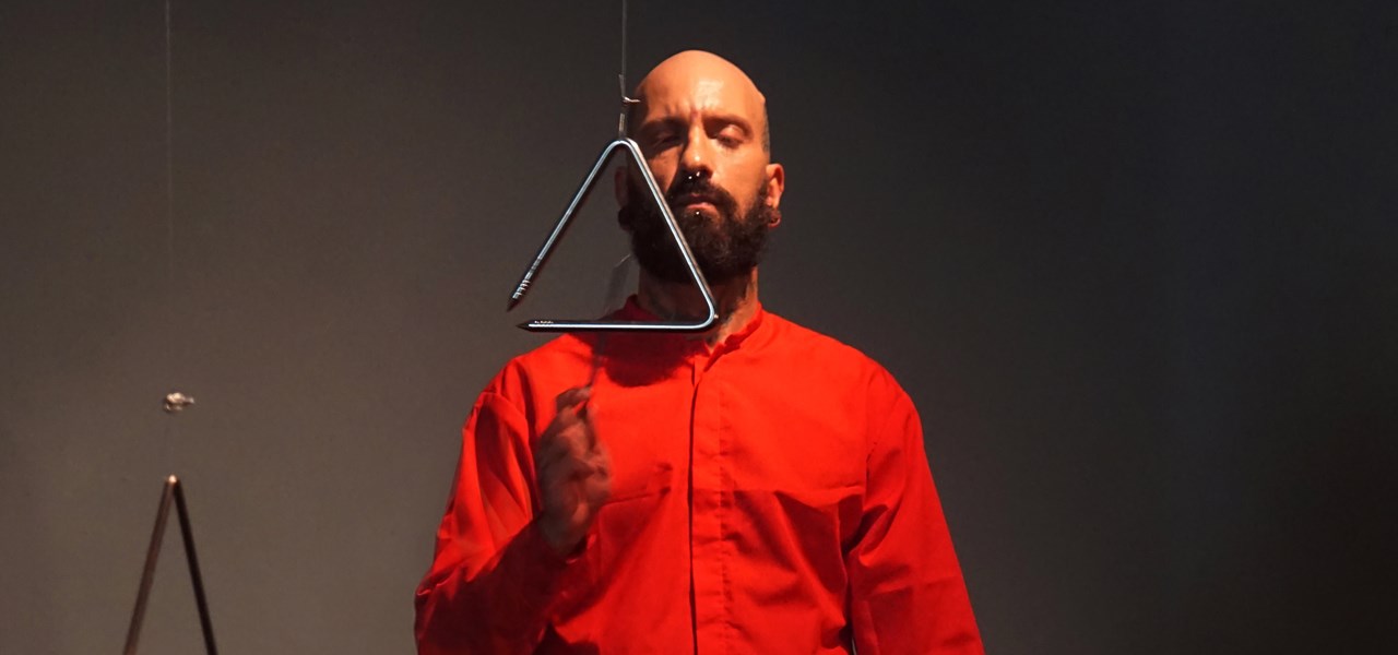 Bald man with full beard striking a triangle, while dressed in a red shirt, against a dark background. 