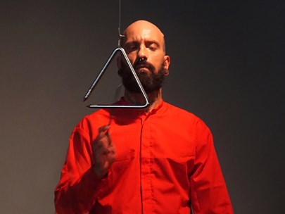 Bald man with full beard striking a triangle, while dressed in a red shirt, against a dark background. 