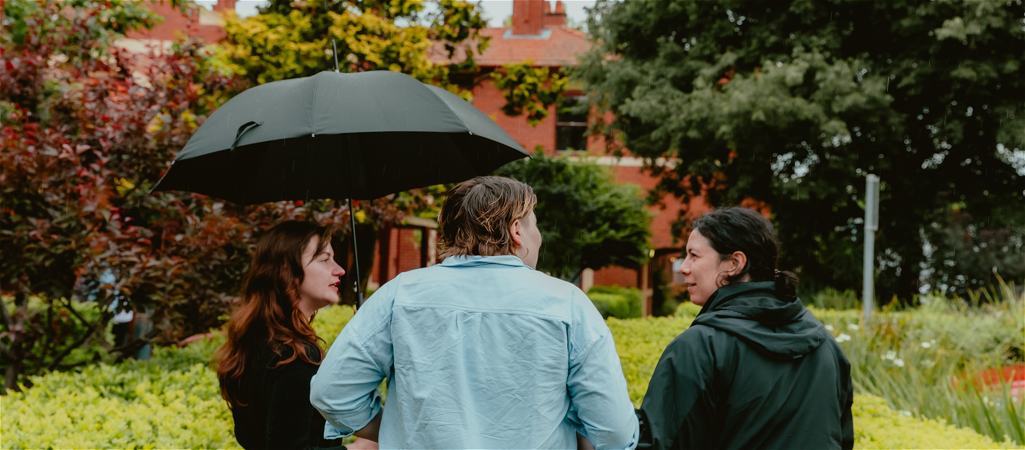 Three people stand talking together while one person is holding an umbrella. They are surrounded by a green garden with a building in the background.