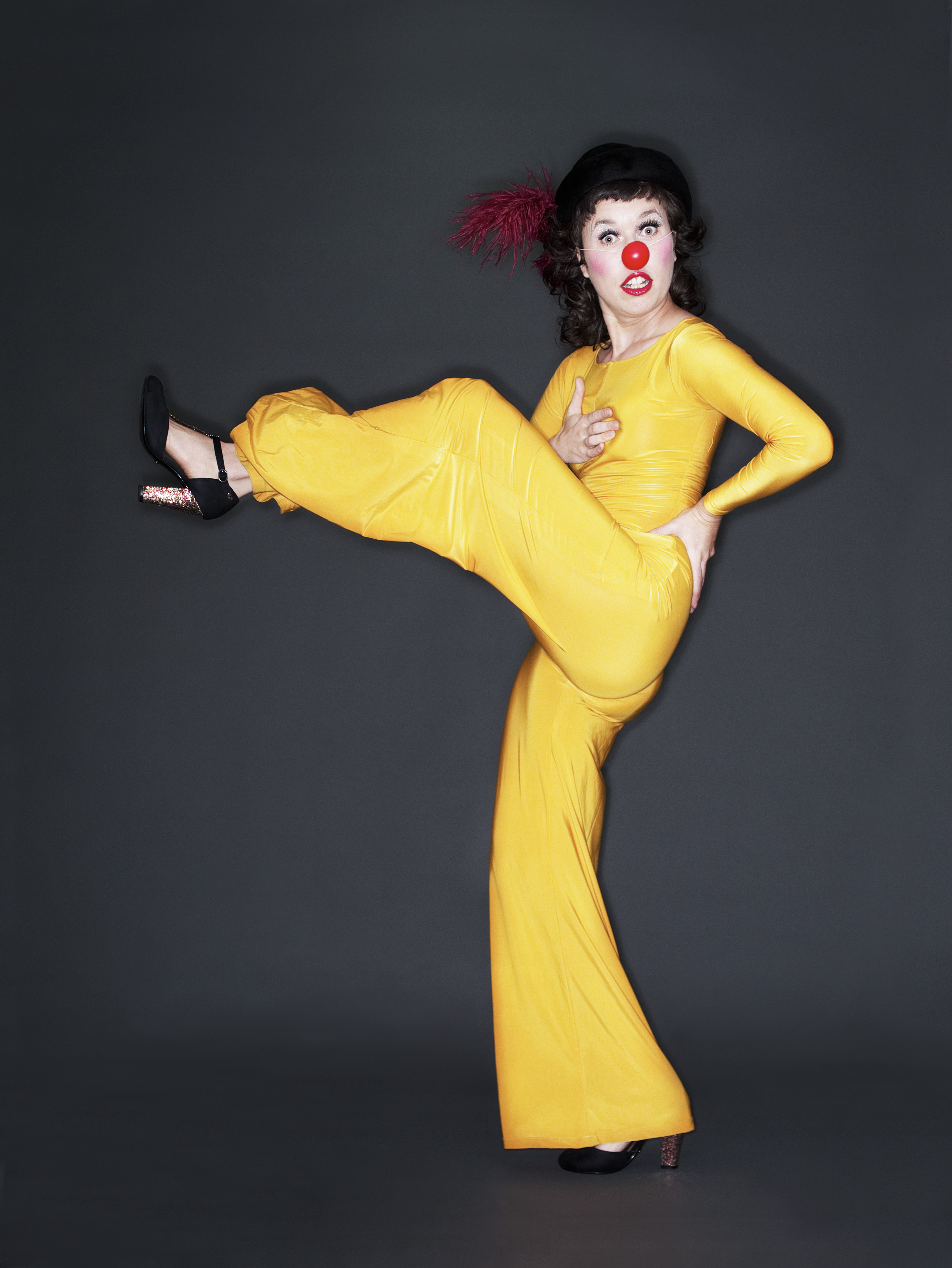Woman in yellow pantsuit and high heels, one leg kicked high, with clown's red nose
