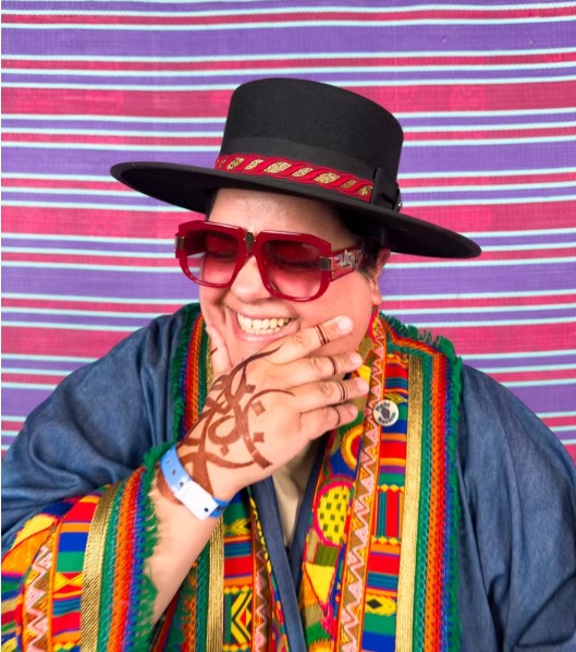 DJ Mz Rizk dressed colourfully against a background of mauve, red and white stripes. 