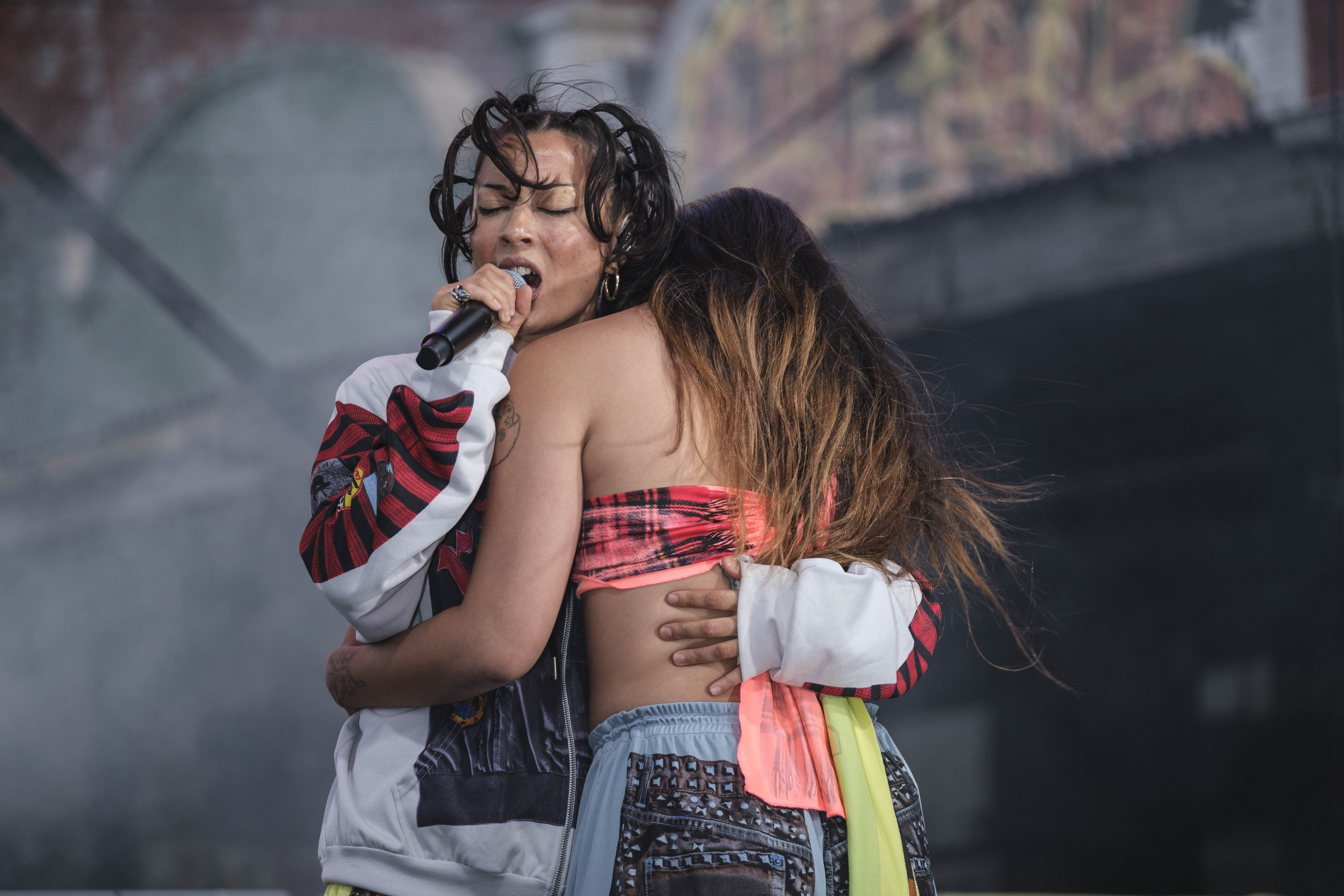 Two female-identifying people hugging. One is singing into a mic with eyes closed. The other has her head on the shoulder of the singer