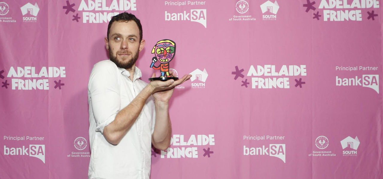 Portrait of Tom holding an award, standing in front of the Adelaide Fringe media wall