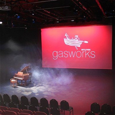 Auditorium showing a few rows of seats then the large stage with a grand piano on it and a large Gasworks sign behind