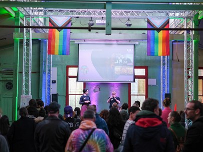 Image of audience looking at a stage with two people on it. There is a projection on a screen above them, with a progress pride flag hanging on either side of the screen.
