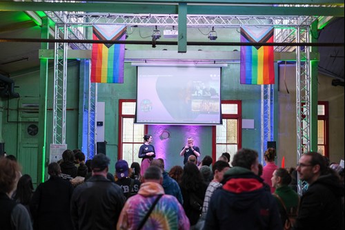 Image of audience looking at a stage with two people on it. There is a projection on a screen above them, with a progress pride flag hanging on either side of the screen.