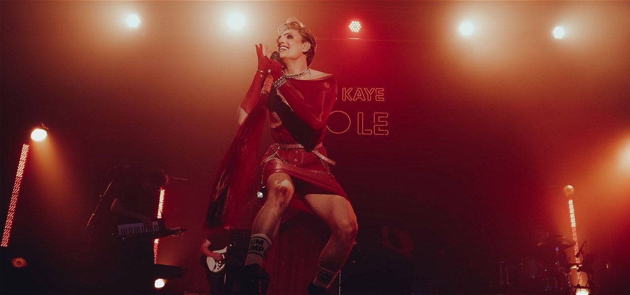 Reuben Kaye on a brightly lit stage wearing all red skirt, blouse and long gloves