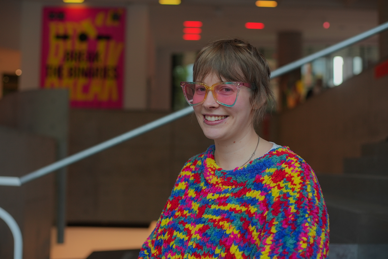 A person wearing a colourful sweater and pink glasses smiling.