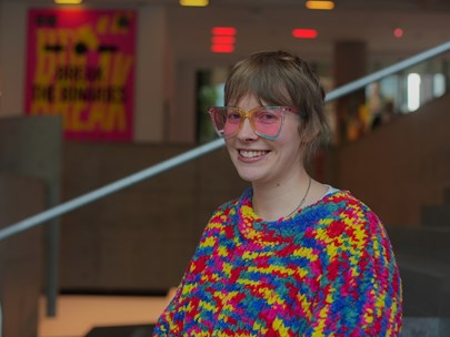 A person wearing a colourful sweater and pink glasses smiling.