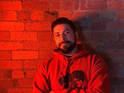 Butch le Butch standing in front of a brick wall wearing a hoodie. Red hues.