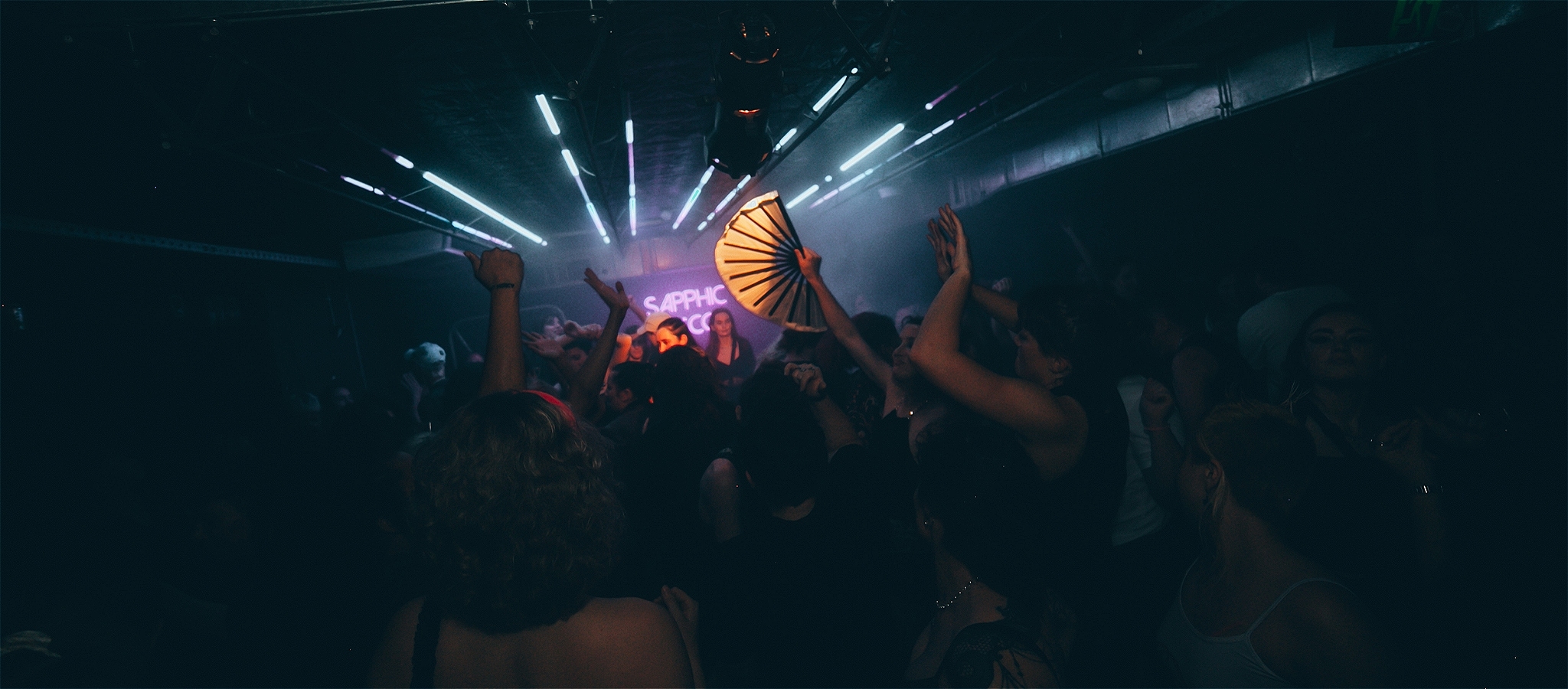 A crowd of people dance, a sign in the background reads Sapphic Disco. A person is waving a fan in the air.
