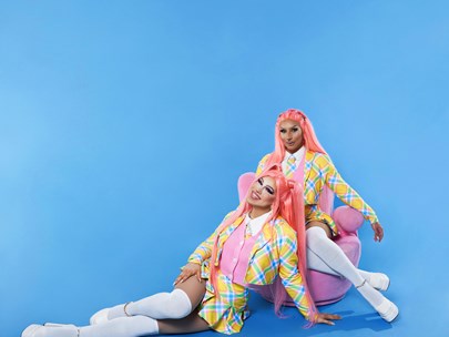 Jawbreakers with a brilliant blue background; one sitting in a pink armchair, the other leaning on them. Both dressed in white stockings, pink blouse, and yellow/blue suit